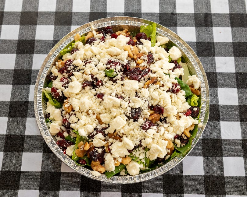 Salad with lettuce, cranberries, walnuts, and feta cheese in a foil container on a checkered tablecloth.