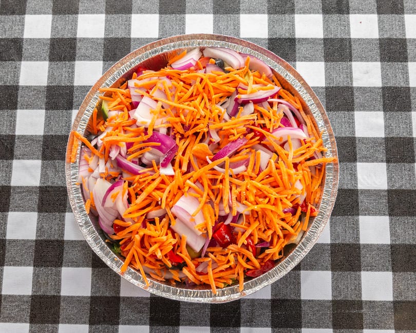 A foil container filled with shredded carrots, red onions, and other vegetables on a checkered tablecloth.