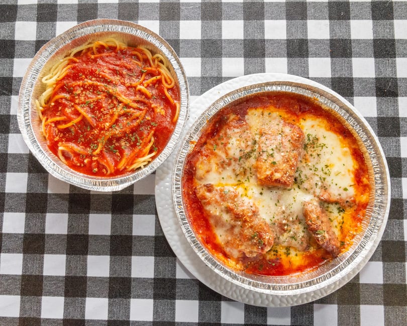 Two foil containers of Italian food, spaghetti with red sauce and chicken parmesan, on a checkered tablecloth.
