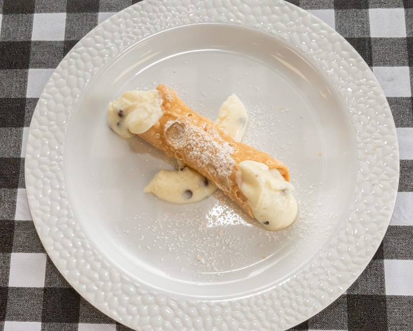 A single cannoli filled with cream and chocolate chips, dusted with powdered sugar on a white plate.