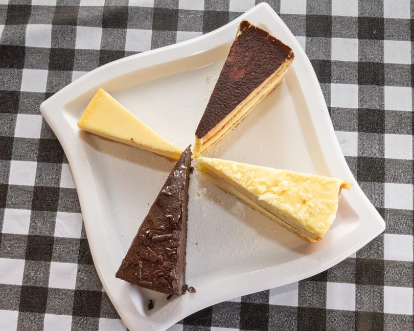 Four different dessert slices, including chocolate and cheesecake, arranged on a white plate over a checkered tablecloth.