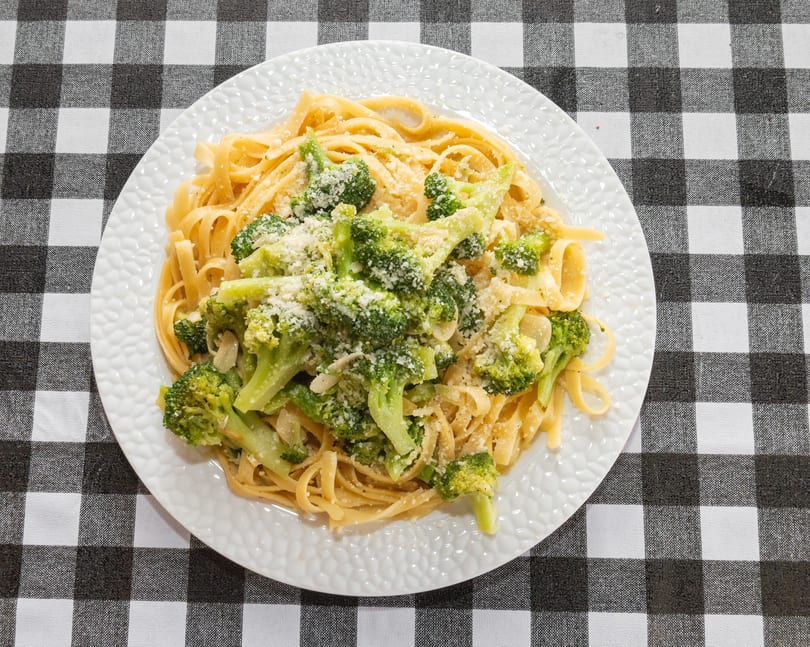Fettuccine pasta with broccoli florets and grated Parmesan cheese on a white plate, on a checkered tablecloth.